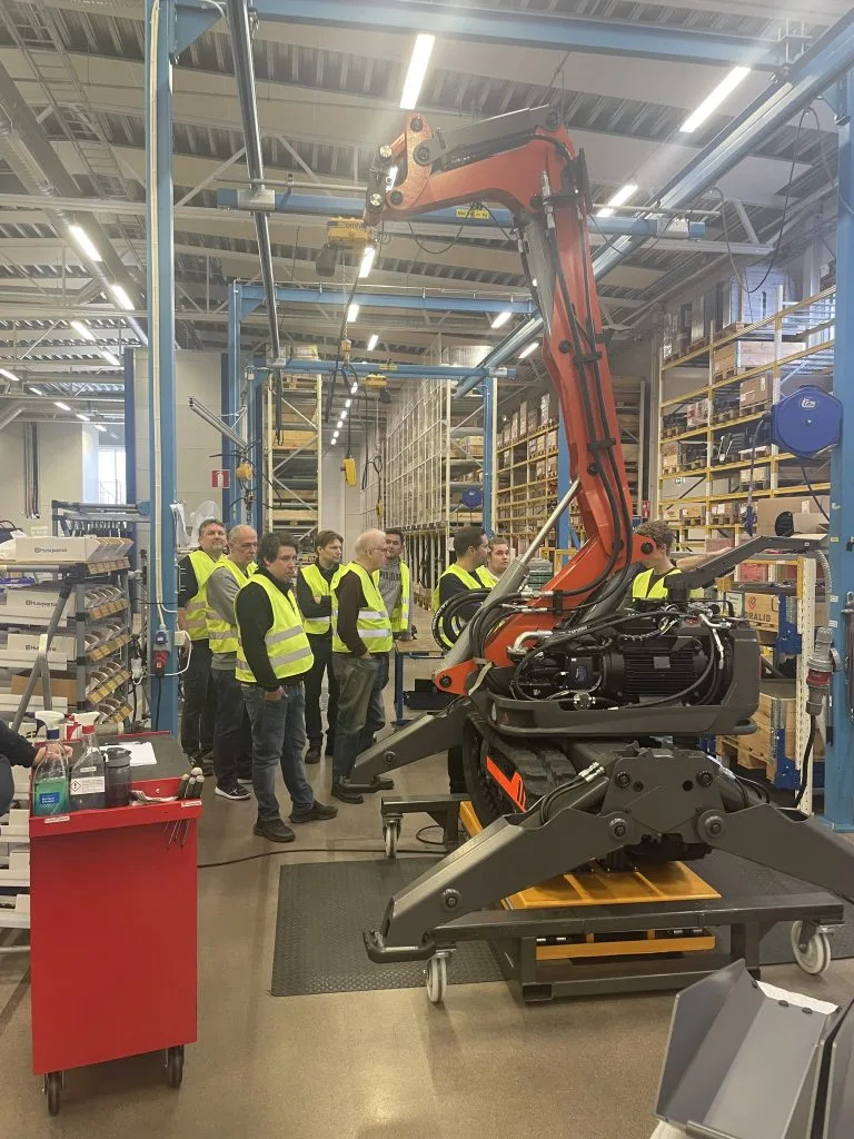 Besökare i gula västar framför en orange robotarm i en produktionslokal